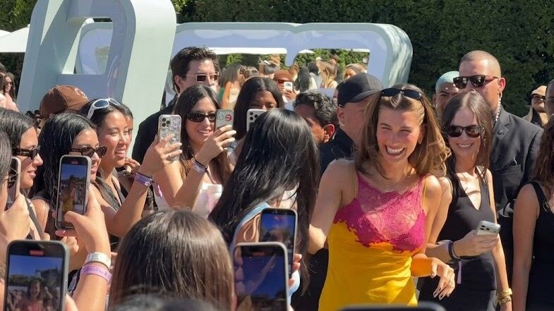 Hailey Bieber tersenyum dan mengenakan gaun kuning dan merah muda dikelilingi oleh penggemar yang mengambil foto di luar ruangan.