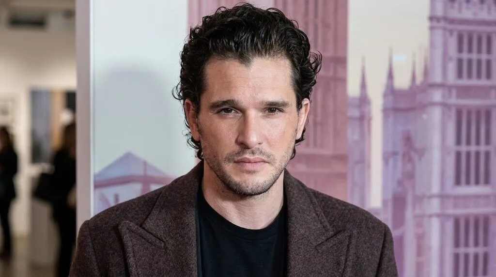 Kit Harington mengenakan mantel coklat dan kemeja hitam, berdiri di dalam ruangan dengan latar belakang kabur.