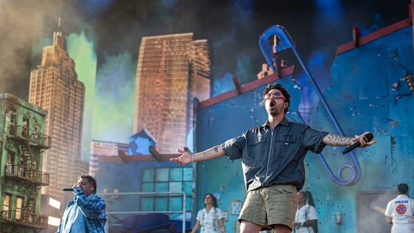 Dua penampil pria bernyanyi di atas panggung dengan latar belakang pemandangan kota dan penari berpakaian putih.