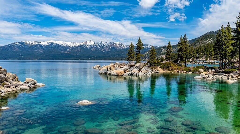 Danau biru kehijauan yang jernih dengan garis pantai berbatu dan pepohonan pinus, pegunungan yang tertutup salju di bawah langit sebagian berawan.