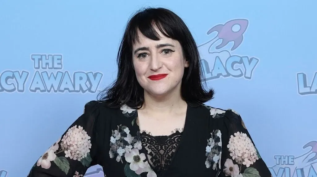 Wanita berambut hitam dan lipstik merah mengenakan gaun bermotif bunga hitam di latar belakang acara The Lacy Awards