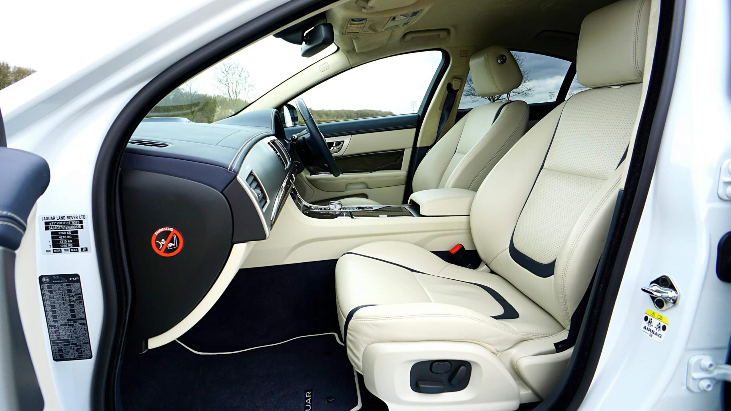 Interior depan Jaguar XF dengan jok kulit krem ​​​​dan trim dashboard hitam terlihat melalui pintu pengemudi yang terbuka.