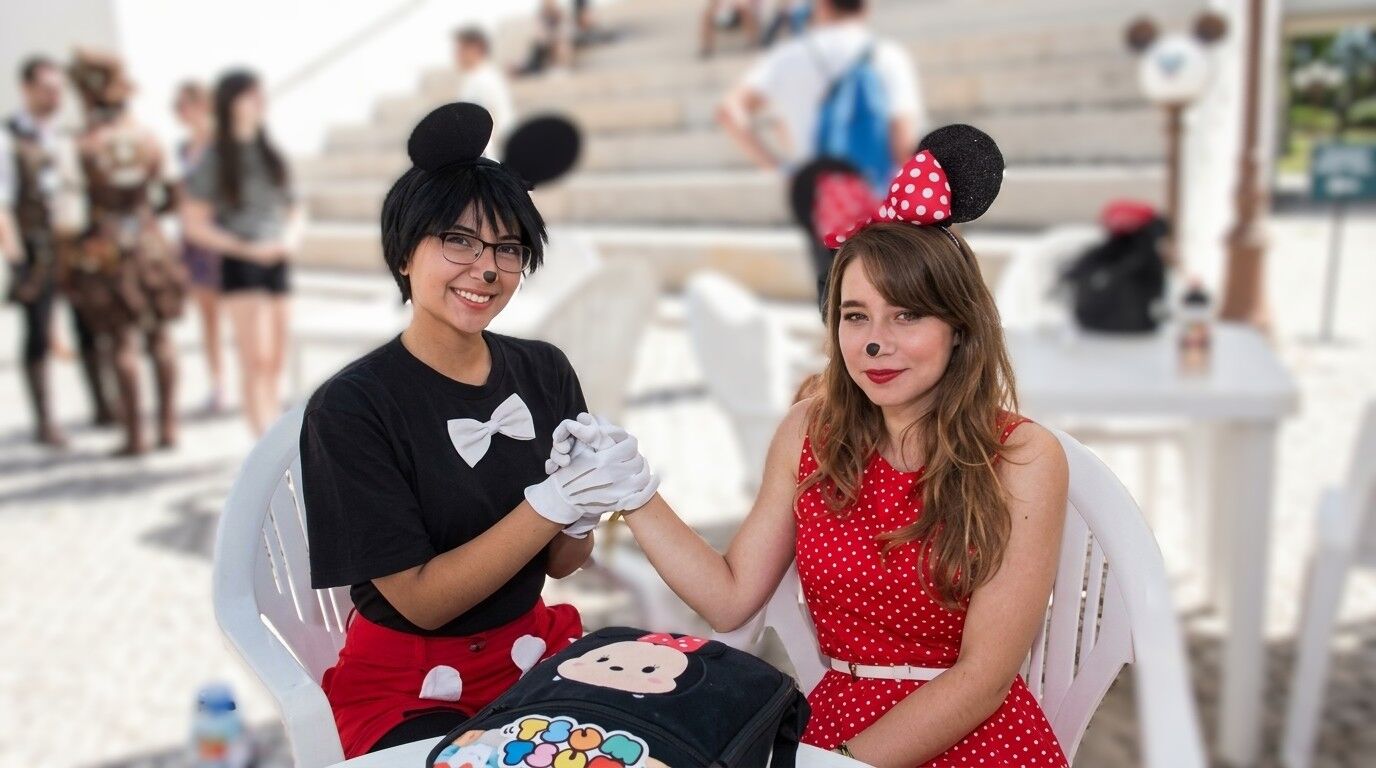 Dua wanita berpakaian Mickey dan Minnie Mouse berpegangan tangan dan tersenyum sambil duduk di luar ruangan