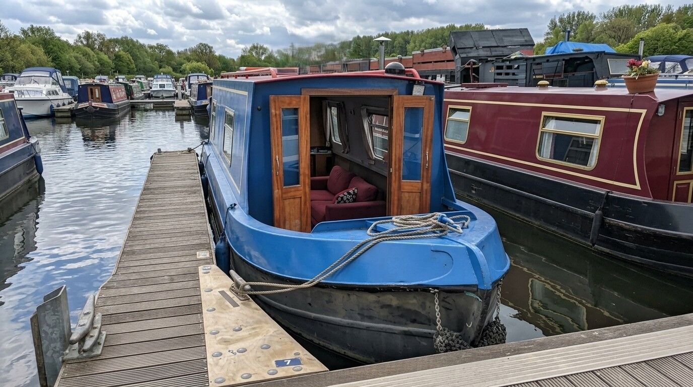 Perahu sempit biru dengan pintu kayu terbuka memperlihatkan sofa merah, berlabuh di marina dengan perahu dan pepohonan lain di latar belakang
