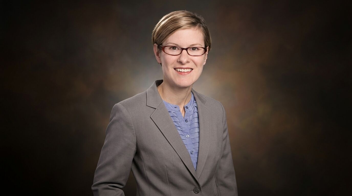 Wanita tersenyum mengenakan kacamata, blazer abu-abu, dan kemeja biru dengan latar belakang studio gelap