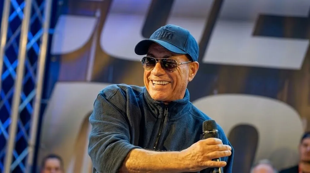 Jean-Claude Van Damme tersenyum dan memegang mikrofon di atas panggung dengan mengenakan kacamata hitam dan topi biru.