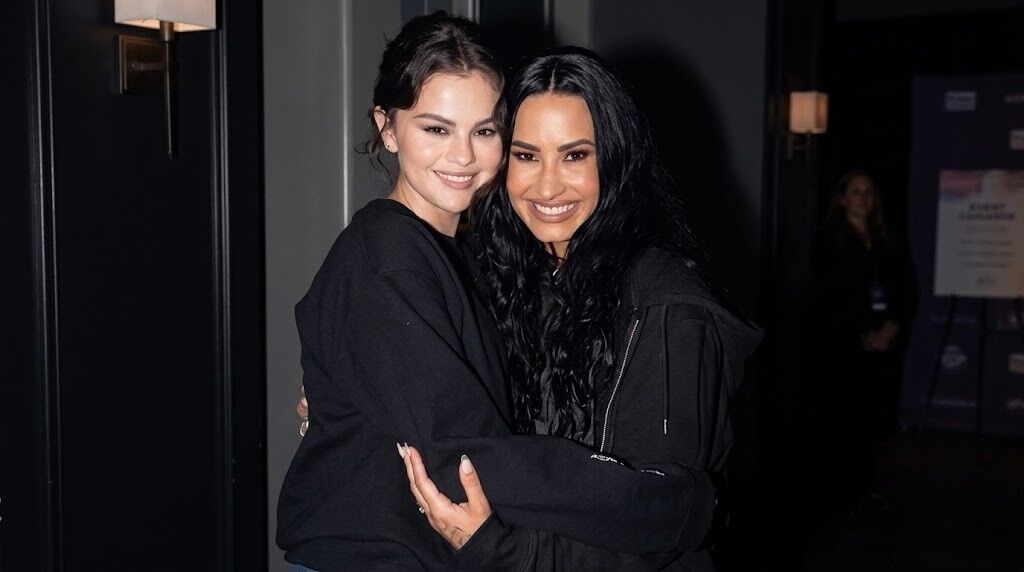 Selena Gomez dan Demi Lovato tersenyum dan berpelukan sambil mengenakan pakaian serba hitam di ruangan yang remang-remang.