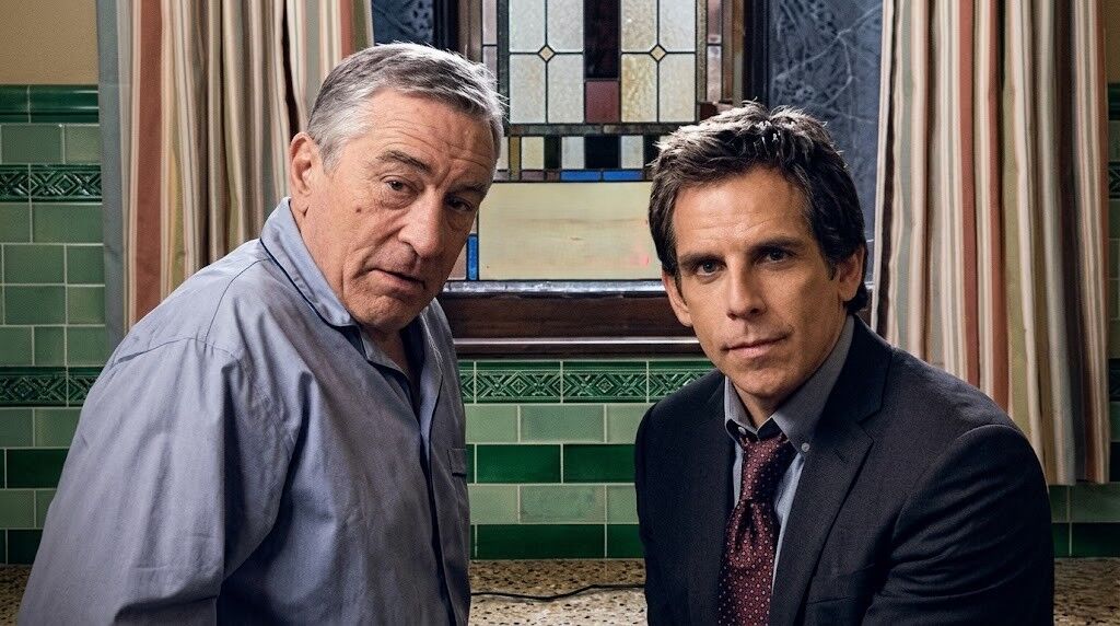 Robert De Niro dan Ben Stiller duduk di dalam ruangan dengan dinding ubin hijau dan jendela kaca patri di belakangnya.