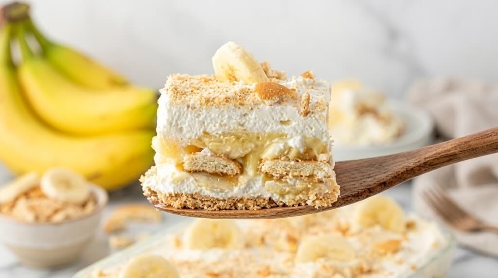 Sepotong puding pisang dengan wafer vanila dan krim kocok di atas spatula kayu dengan pisang di latar belakang