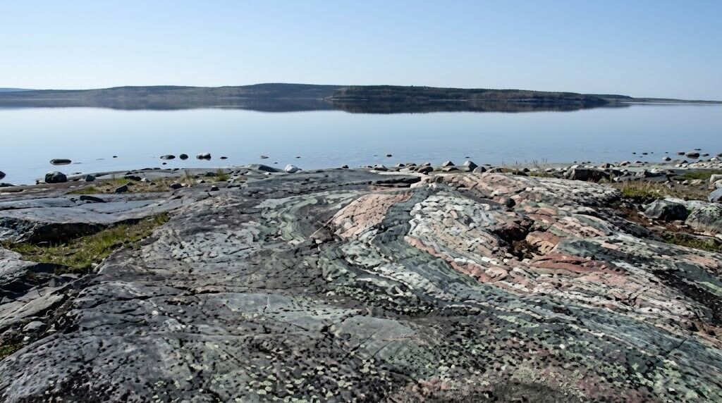 Formasi batuan berlapis warna-warni di tepi danau yang tenang dengan garis pantai berhutan jauh di bawah langit cerah.