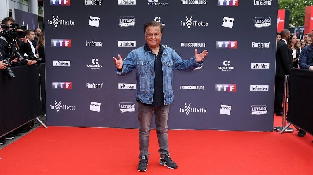 Pria berjaket denim dan celana jeans berpose di karpet merah pada acara dengan latar belakang logo La Villette dan Le Parisien