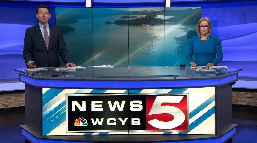 Dua pembawa berita di meja WCYB News 5 dengan latar belakang sungai yang indah di layar studio di belakang mereka.