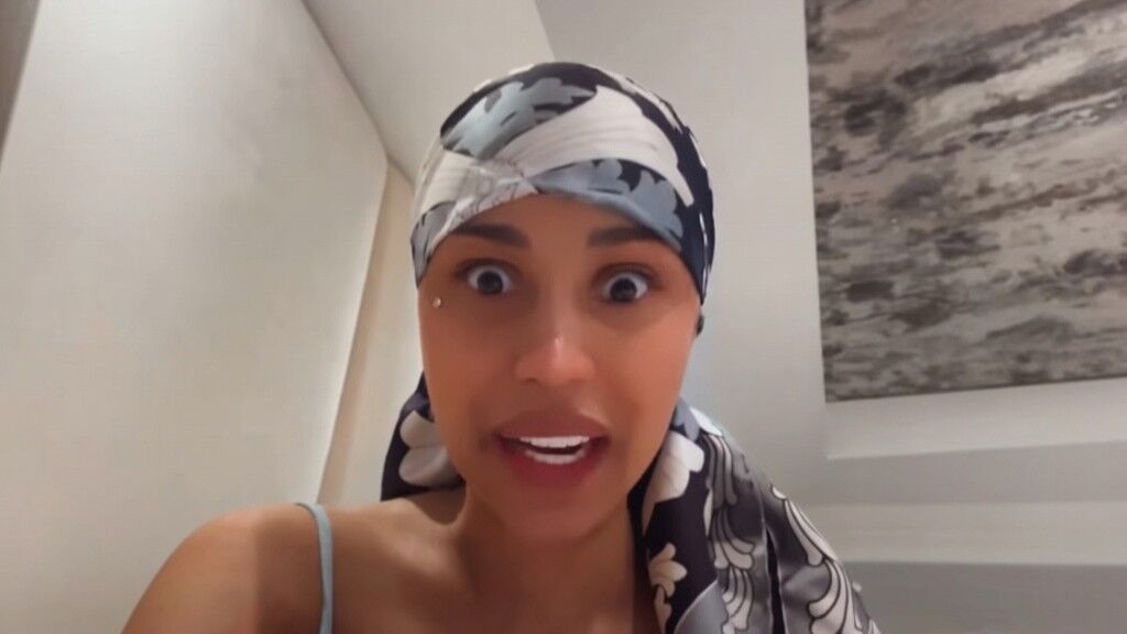 Wanita mengenakan jilbab bermotif bunga hitam putih berbicara dengan mata terbelalak di ruangan berdinding putih dan seni abstrak