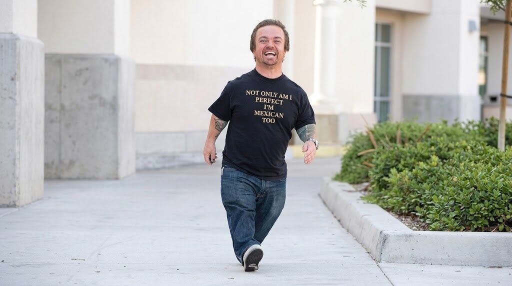 Pria tersenyum dan berjalan di luar ruangan mengenakan kemeja hitam bertuliskan 