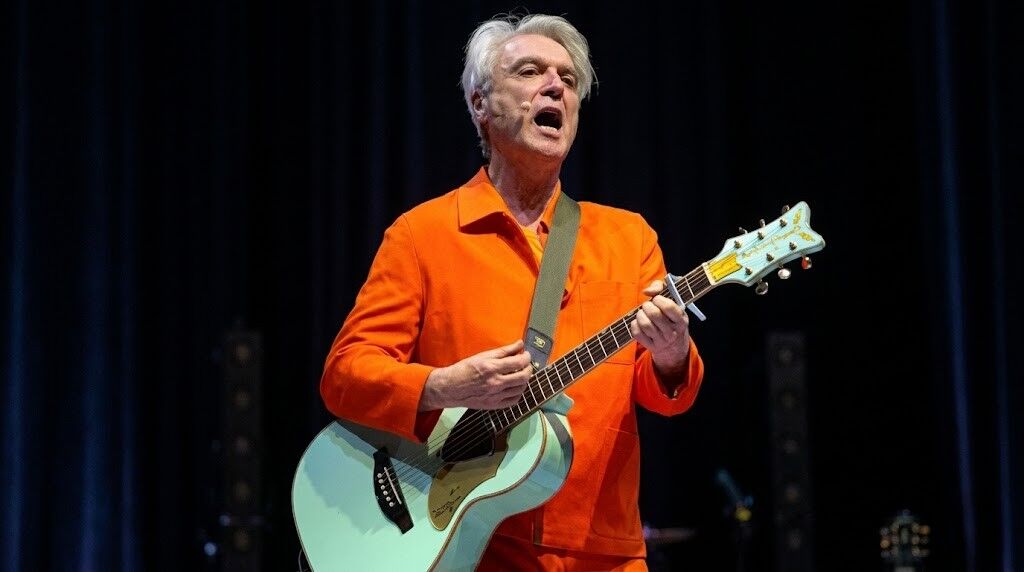 David Byrne tampil di atas panggung dengan mengenakan jaket oranye dan memainkan gitar berwarna biru muda