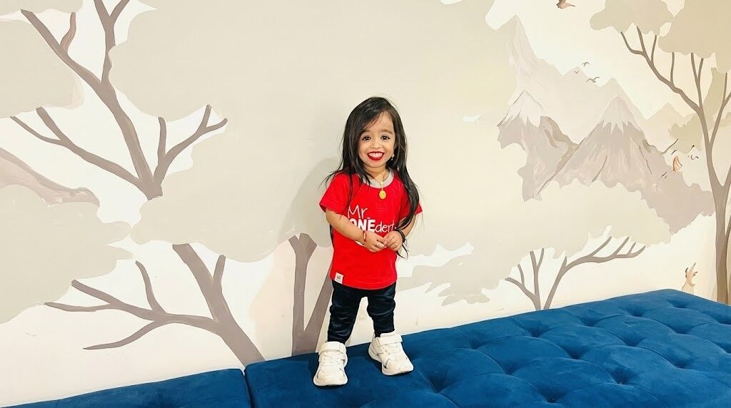 Gadis muda tersenyum dengan kemeja merah dan celana hitam berdiri di sofa biru dengan latar belakang mural pohon dan gunung