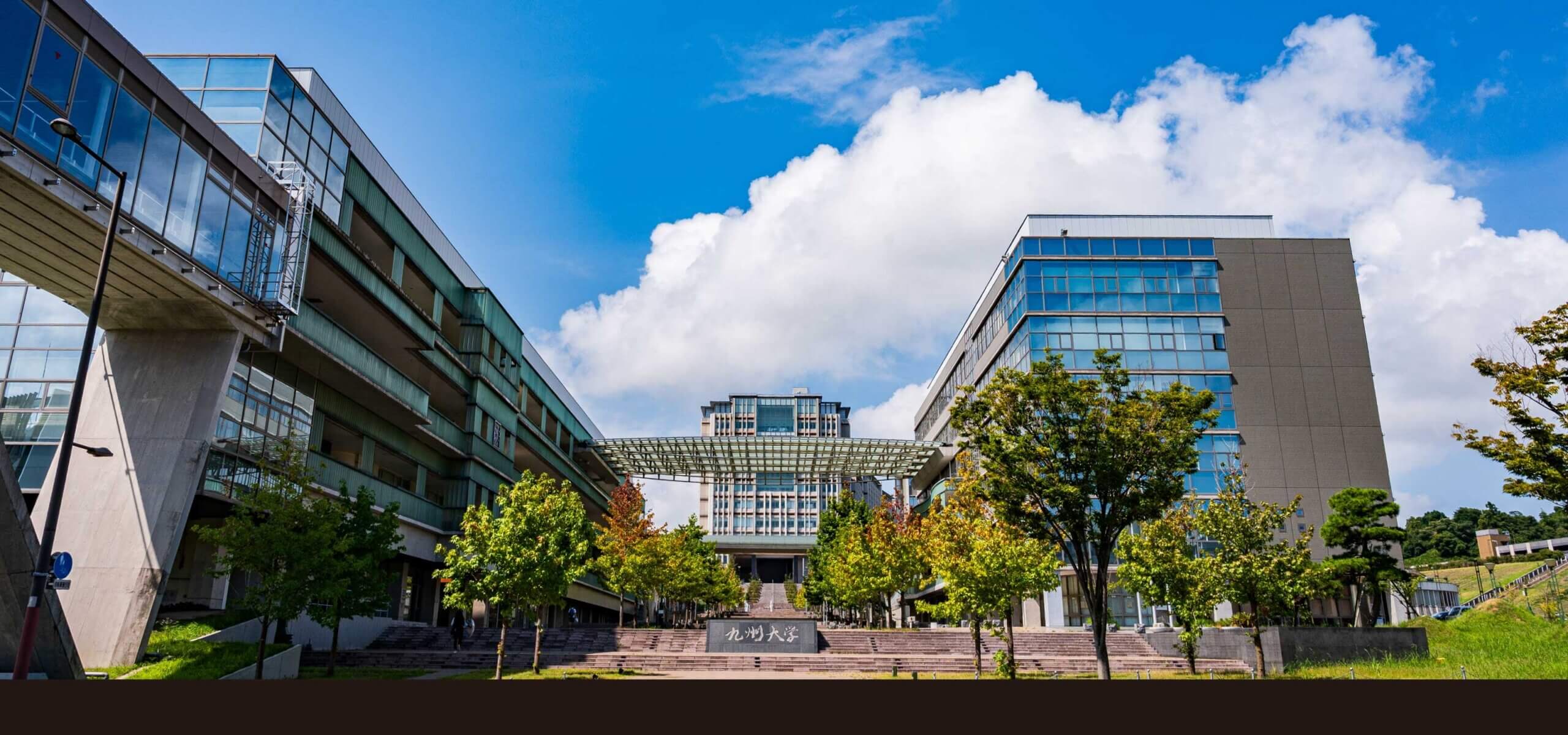 Gedung kampus Universitas Kyushu dengan pepohonan dan langit biru di Fukuoka, Jepang.
