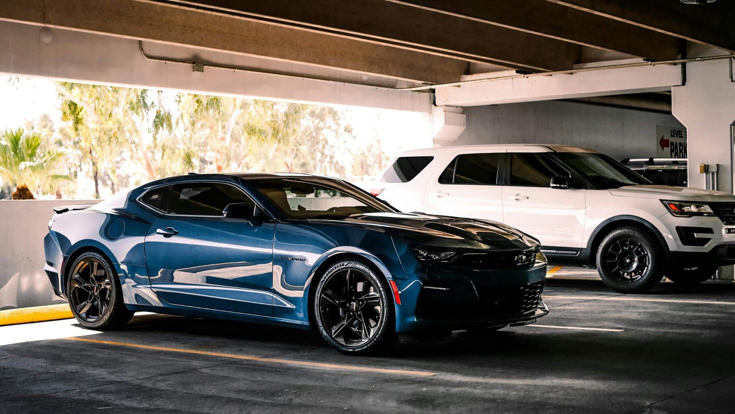 Chevrolet Camaro biru diparkir di samping SUV putih di garasi parkir yang teduh.