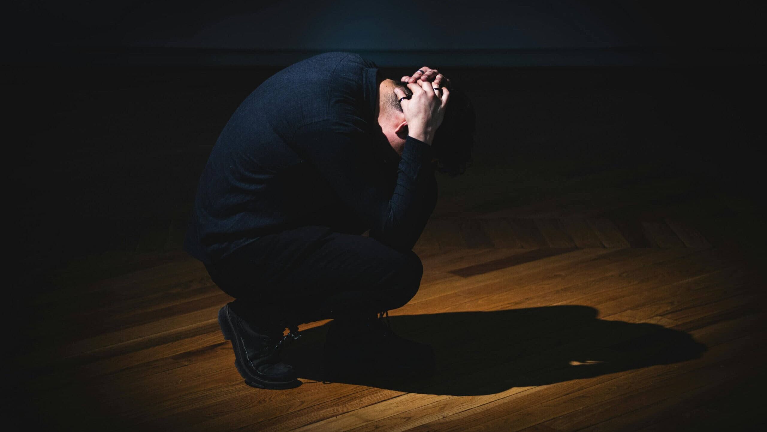 Pria berjongkok di lantai kayu dengan tangan terlipat di kepala dalam cahaya redup