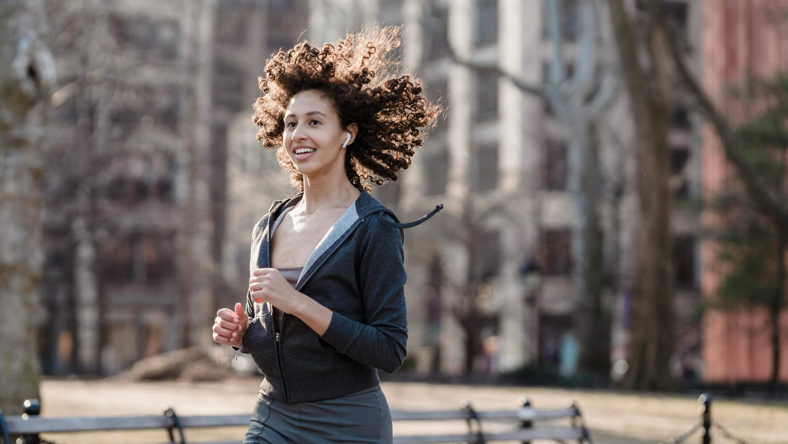 Wanita muda jogging di luar ruangan di taman mengenakan hoodie gelap dan earbud nirkabel