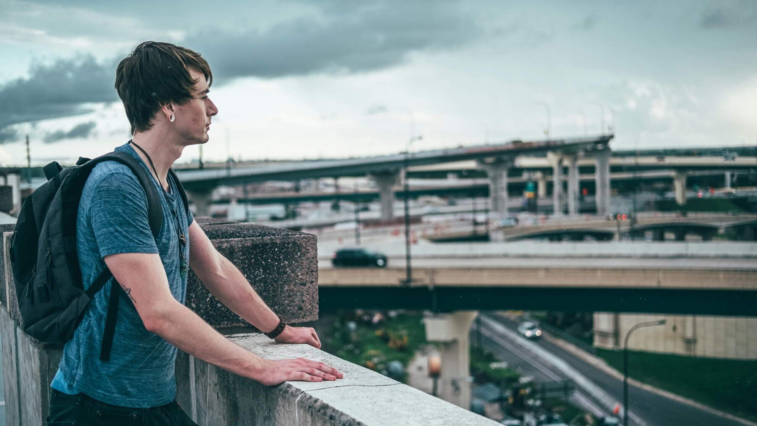 Pemuda dengan ransel bersandar di pagar beton menghadap jembatan layang di bawah langit mendung