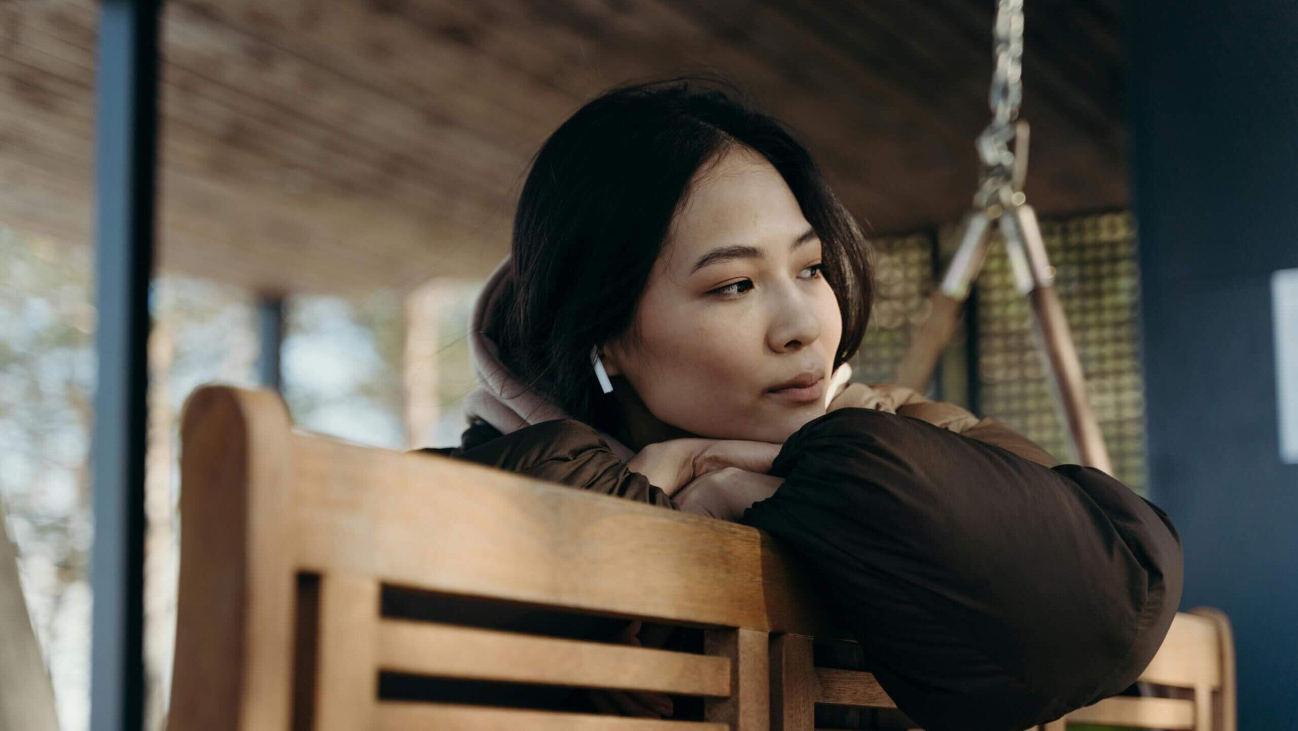 Wanita muda yang mengenakan AirPods sedang beristirahat di bangku kayu, memandang ke samping sambil berpikir.