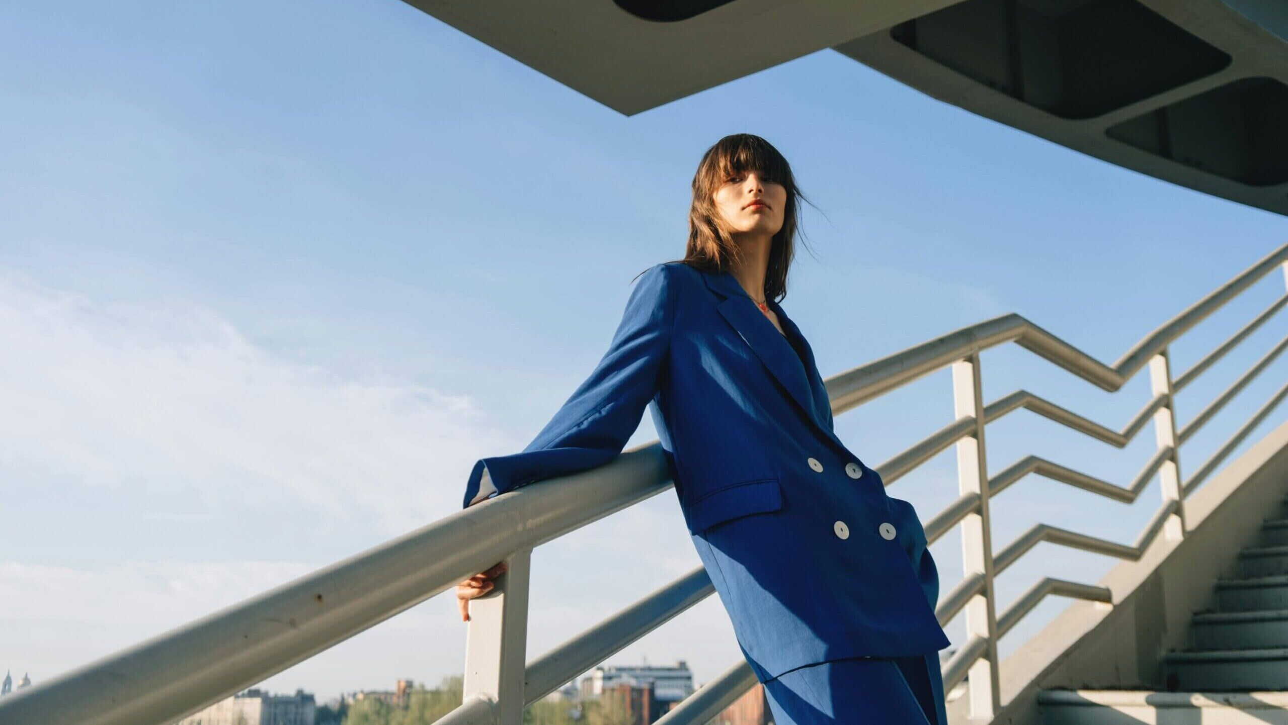 Wanita berjas double-breasted biru bersandar di pagar di bawah struktur modern dengan latar langit cerah