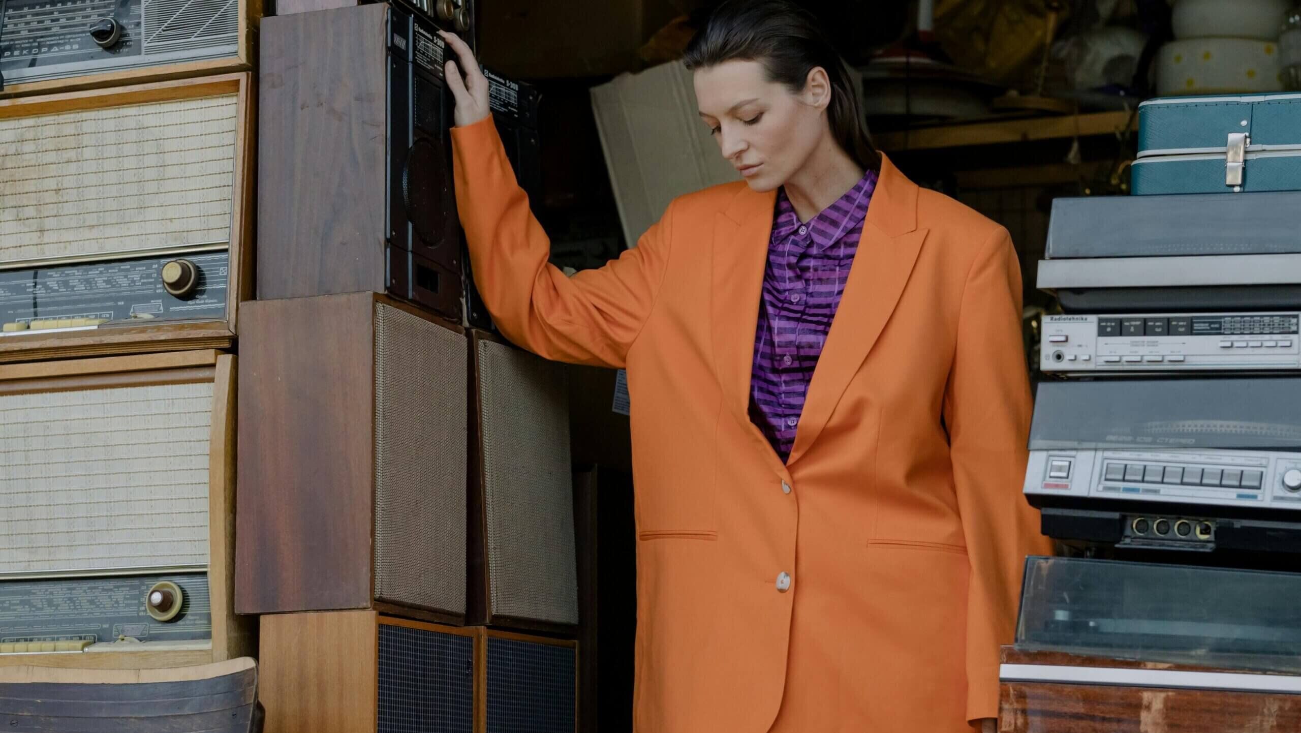 Wanita berjas oranye dan kemeja ungu berdiri di antara radio antik dan perlengkapan audio.