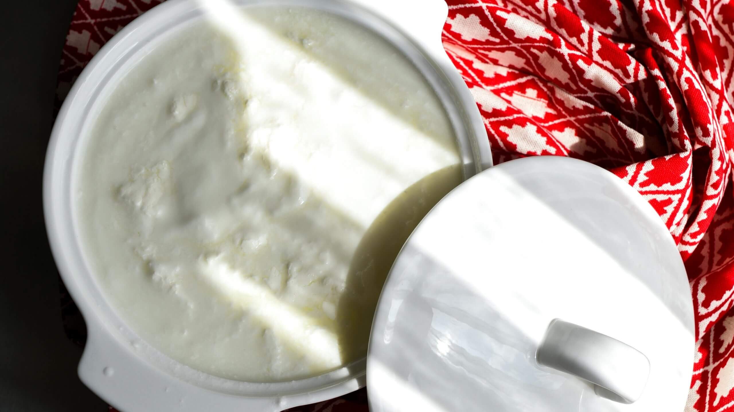 Panci keramik putih dengan tutup terbuka sebagian memperlihatkan bagian dalam yogurt kental di atas kain bermotif merah.