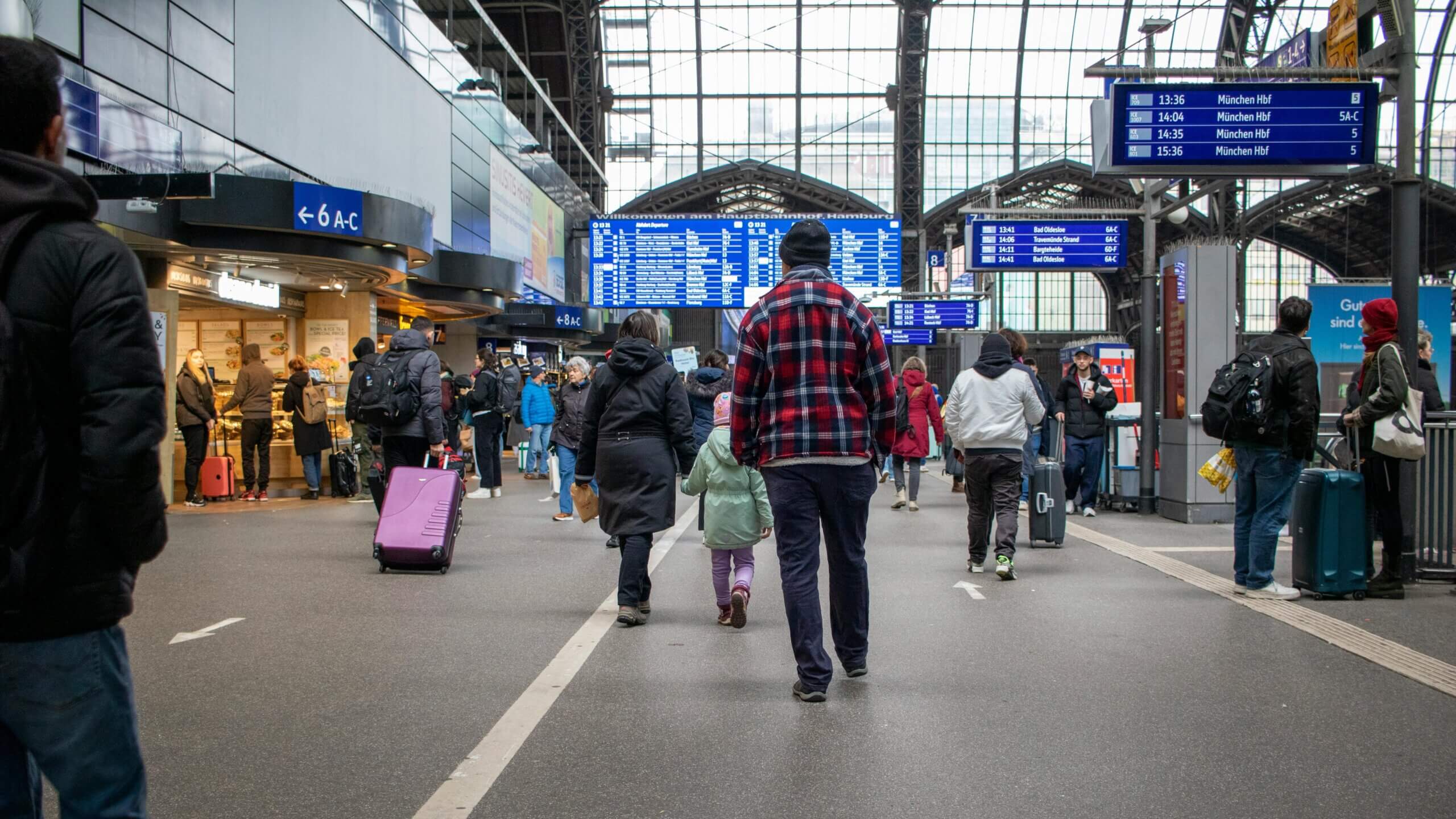 Wisatawan berjalan dan menarik barang bawaan di dalam stasiun kereta Hamburg Hauptbahnhof dengan papan keberangkatan di atasnya.