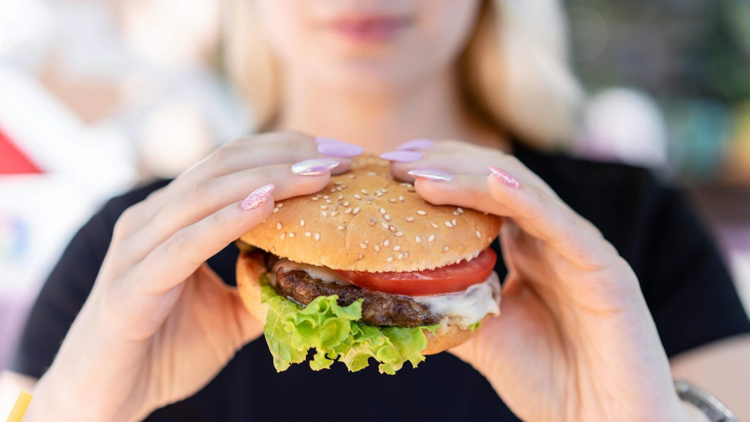 Orang dengan kuku merah muda berkilauan memegang burger keju dengan selada dan tomat dalam roti wijen