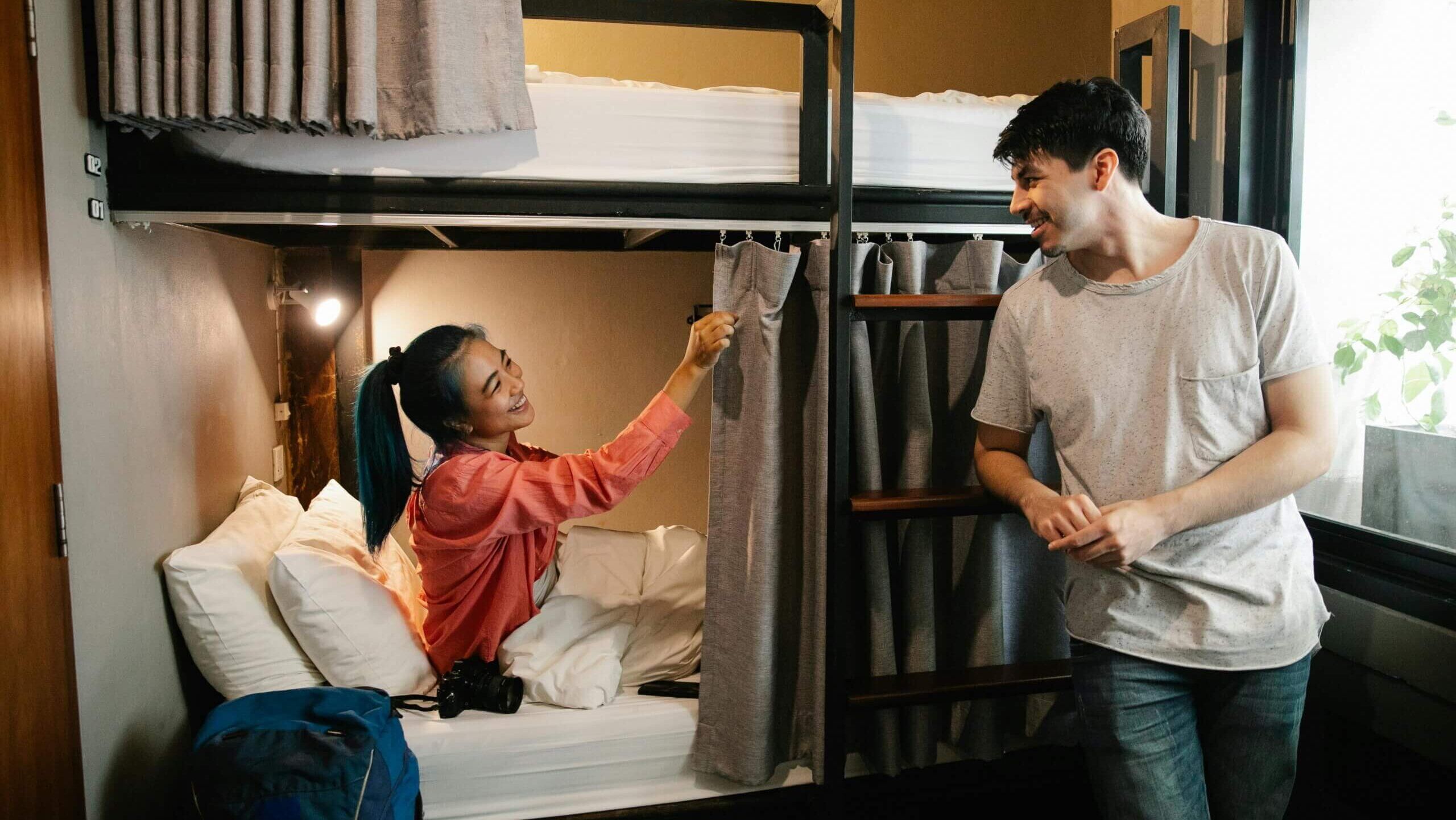 Wanita tersenyum duduk di tempat tidur bawah menarik tirai sementara pria berdiri di dekatnya dengan pakaian kasual di kamar asrama