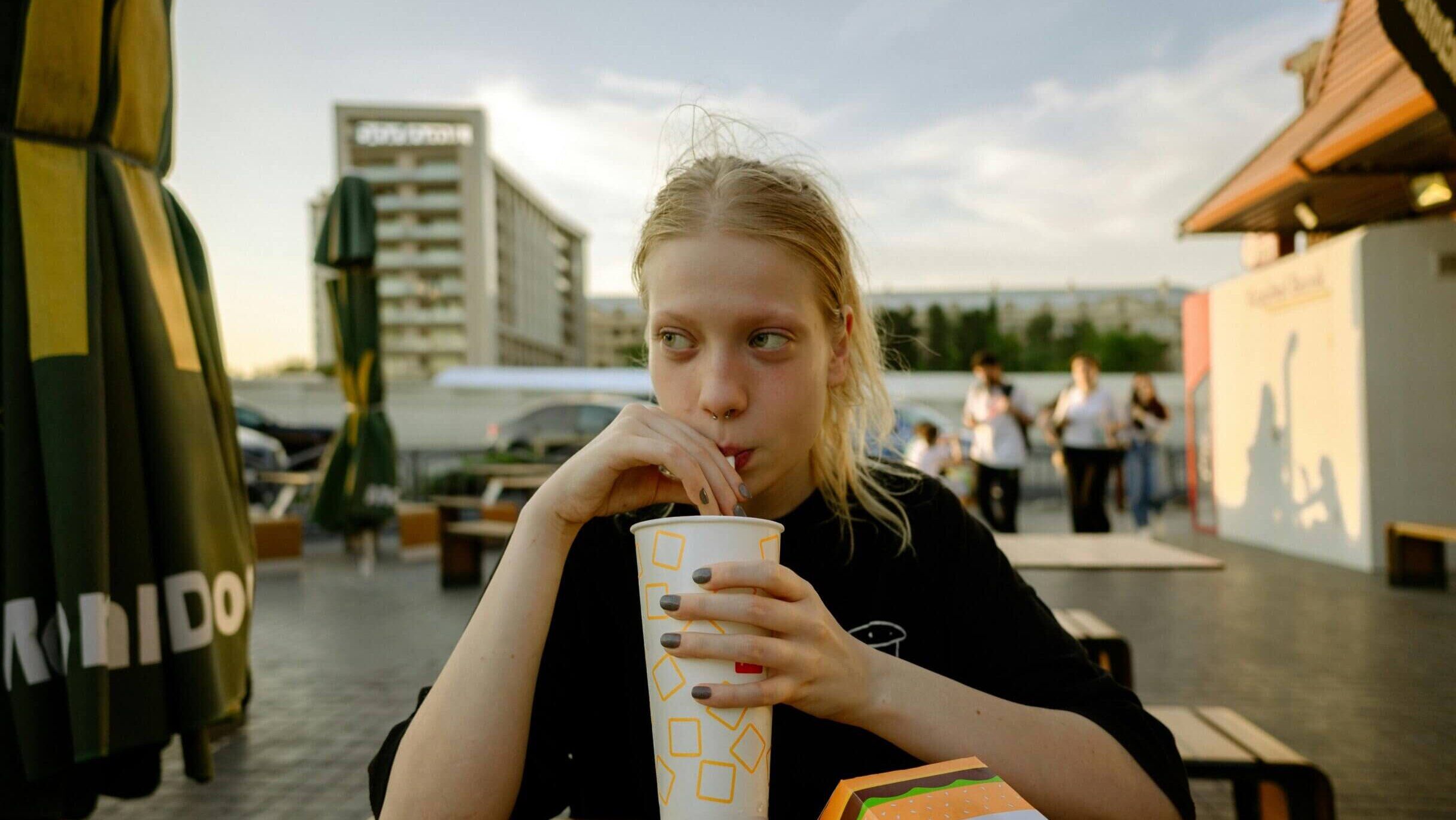 Wanita muda dengan tindik septum minum dari cangkir di area tempat duduk makanan cepat saji luar ruangan dengan kotak Big Mac di dekatnya