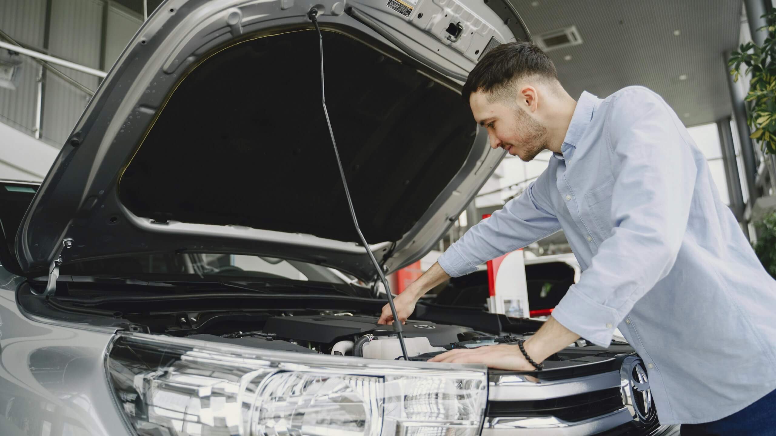 Seorang pria memeriksa mesin di bawah kap kendaraan Toyota berwarna perak di ruang pamer atau garasi.
