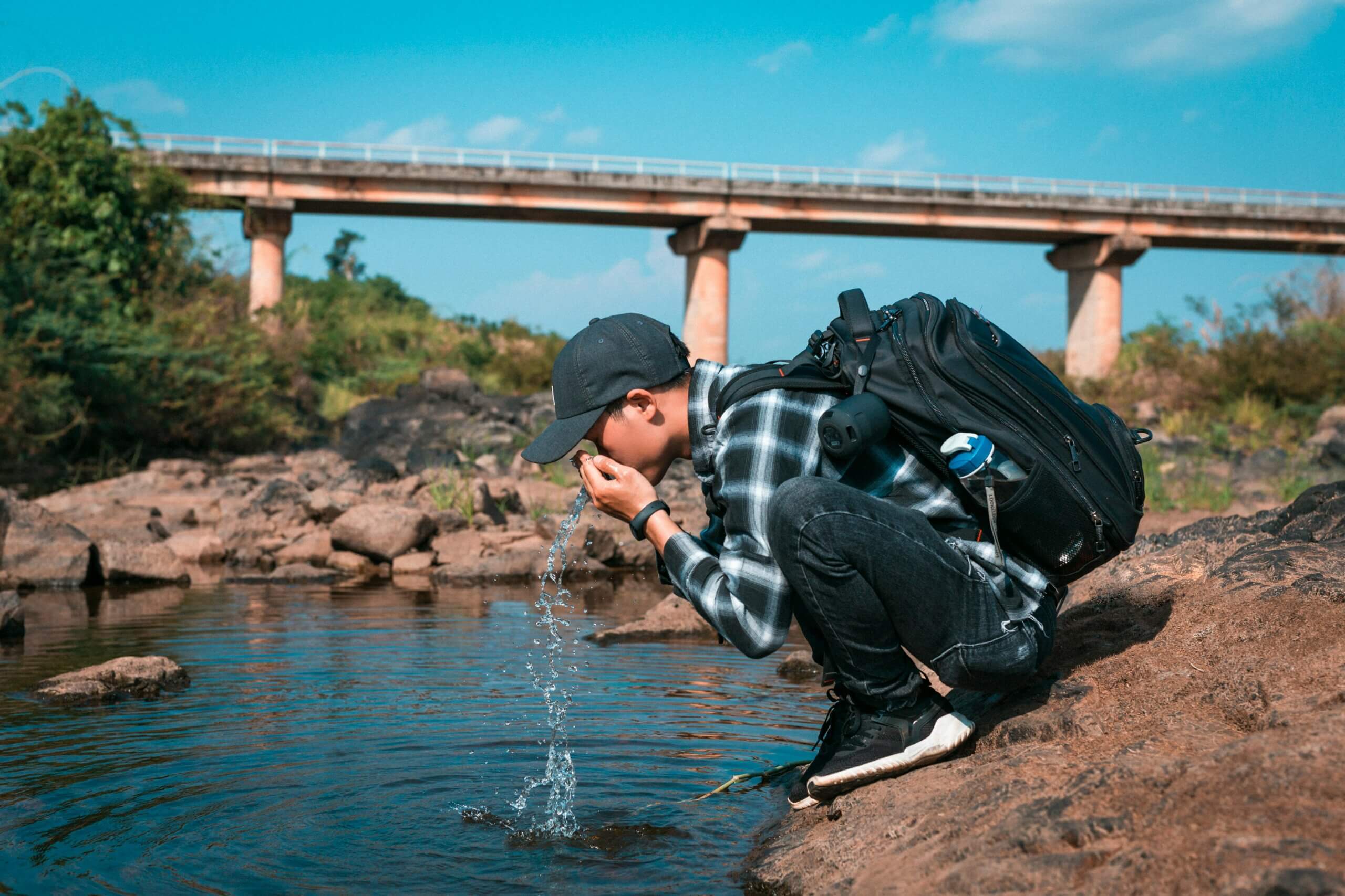 Orang yang memakai topi hitam dan ransel berjongkok di tepi sungai sambil minum air dengan latar belakang jembatan.