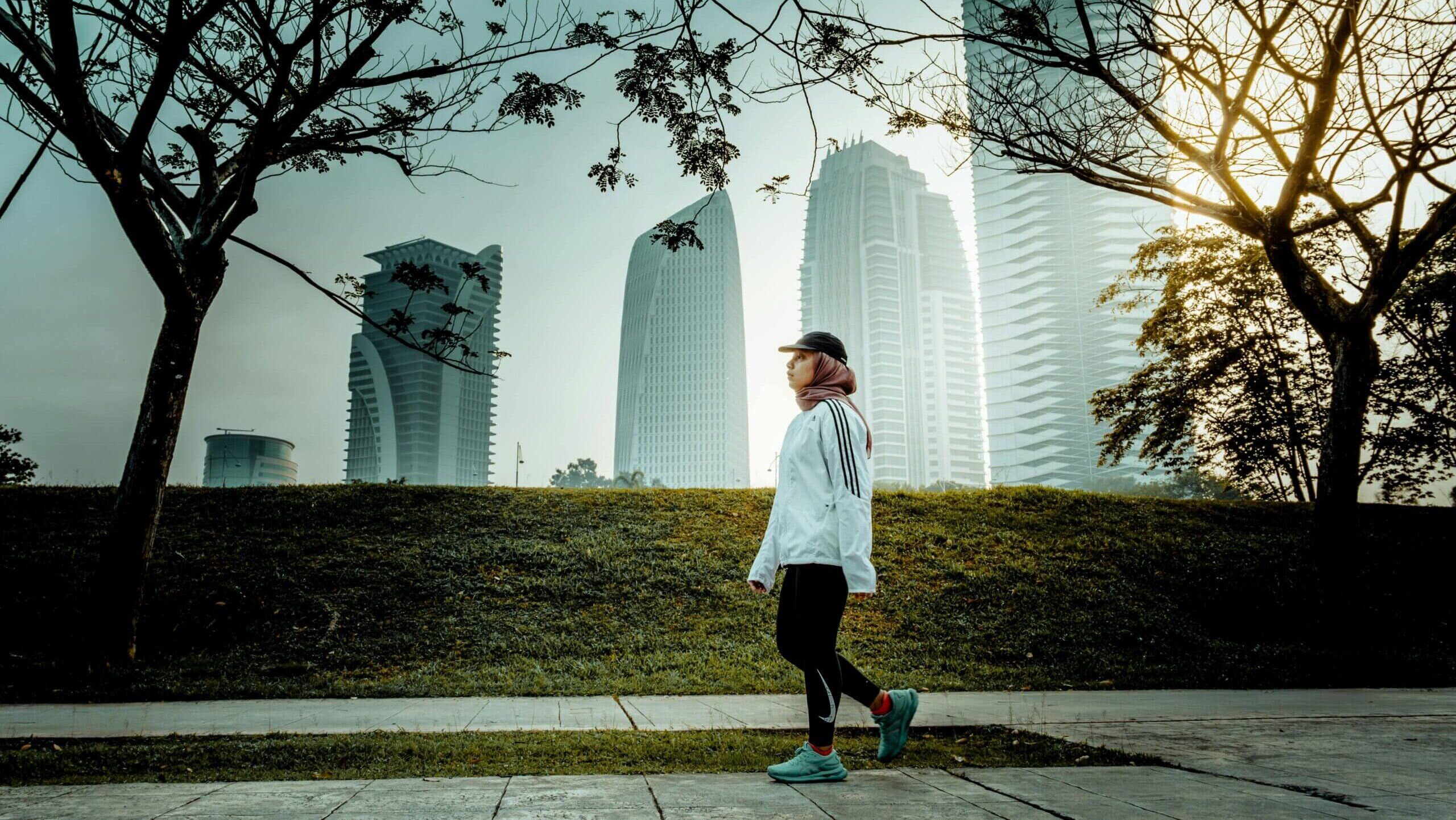 Wanita berjaket Adidas putih dan berhijab berjalan di taman dengan latar belakang gedung pencakar langit modern saat matahari terbit.