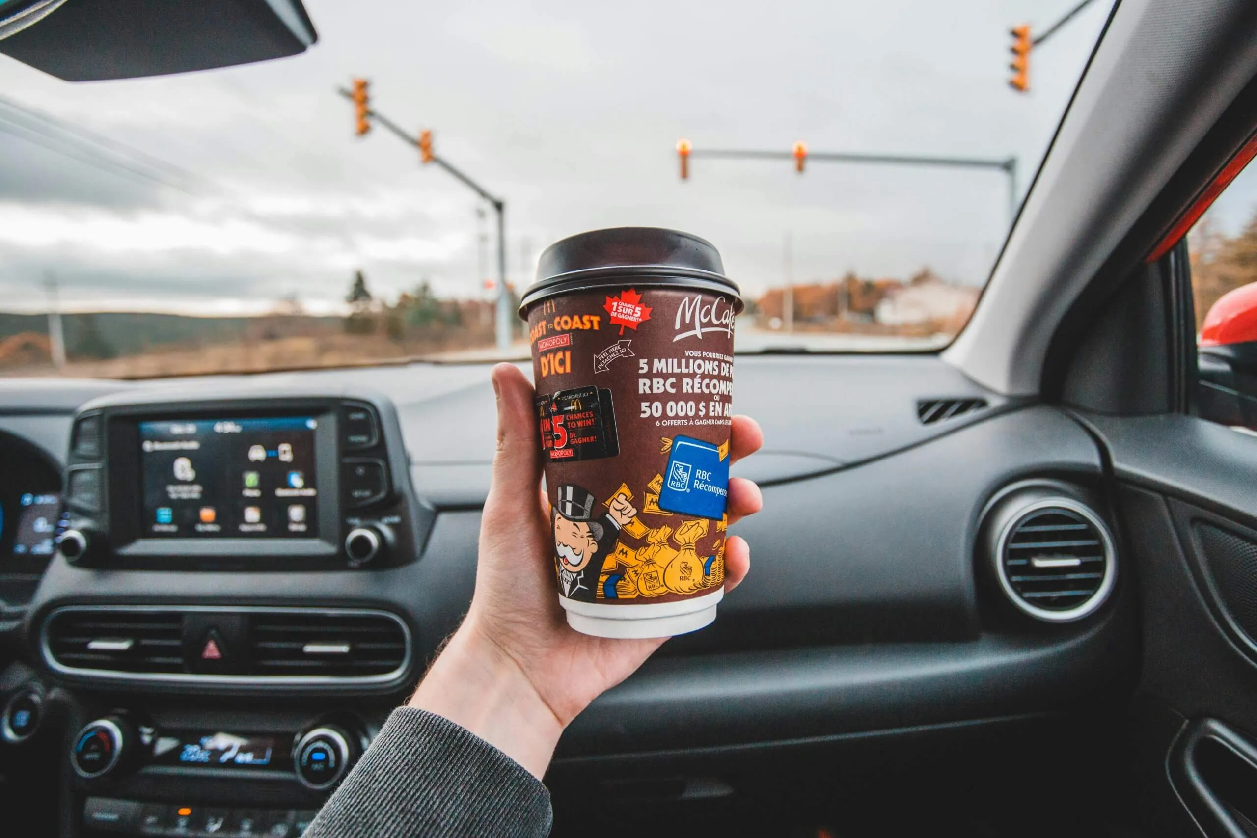 Tangan memegang cangkir kopi McCafé di dalam mobil dengan dashboard dan lampu lalu lintas di latar belakang.