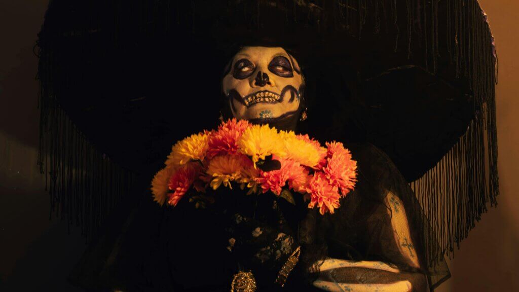 Orang dengan cat wajah tengkorak mengenakan topi hitam besar dan memegang bunga marigold oranye dan kuning untuk perayaan Día de los Muertos