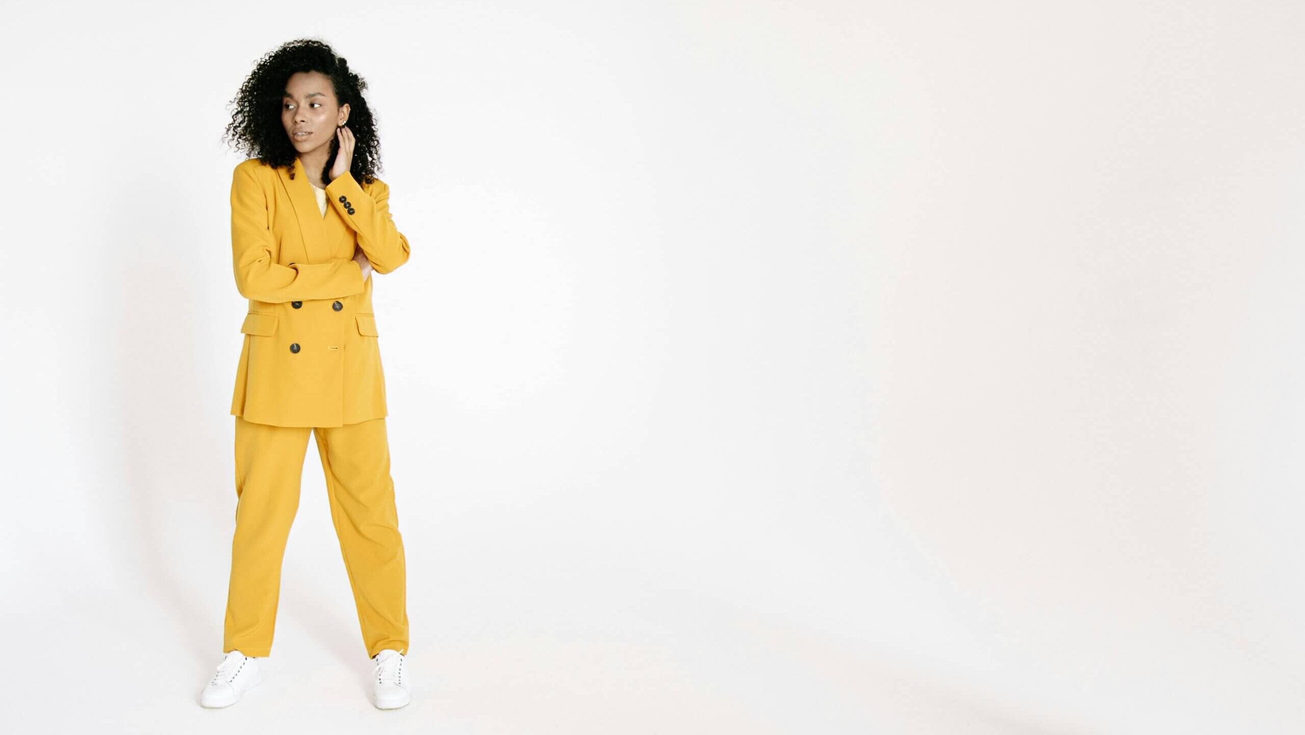 Wanita berambut keriting mengenakan setelan kuning mustard dan sepatu kets putih berdiri dengan latar belakang putih