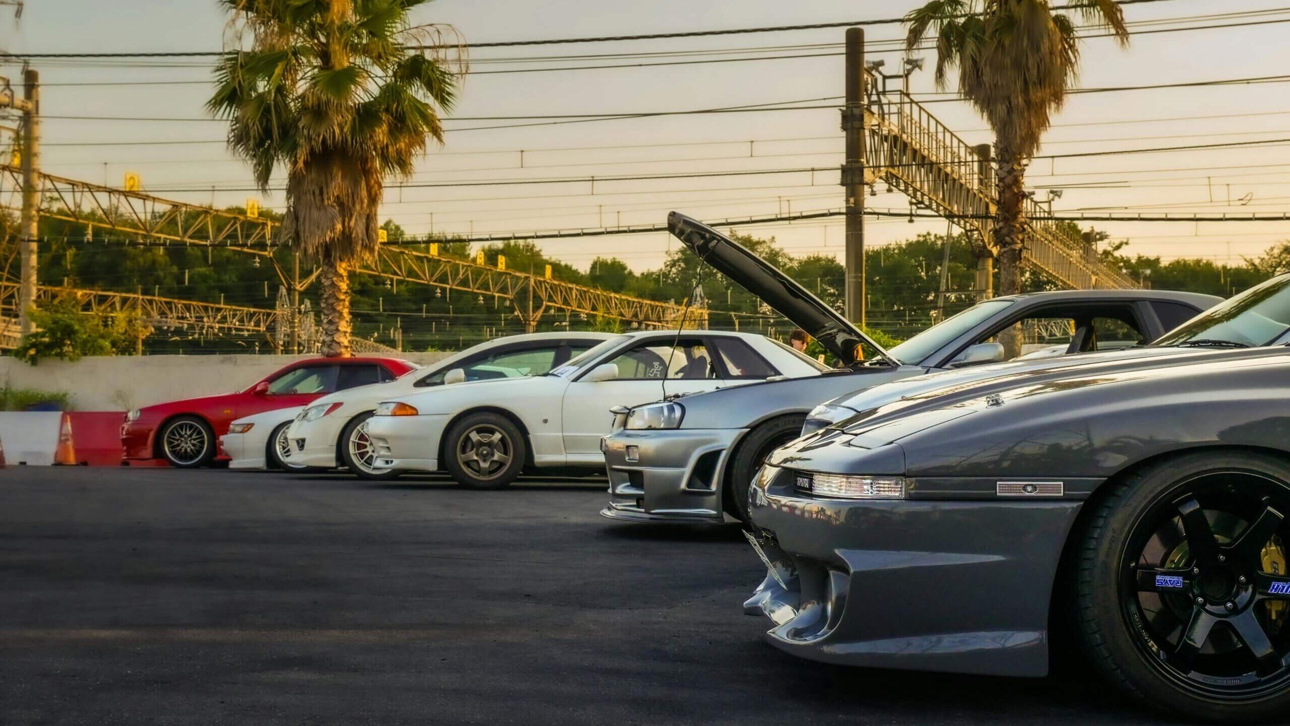 Jajaran mobil sport yang dimodifikasi termasuk model Nissan Skyline diparkir di dekat pohon palem saat matahari terbenam
