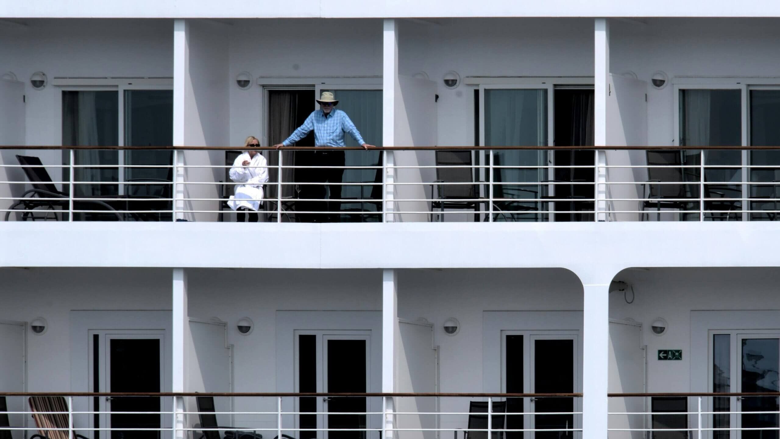 Dua orang di balkon kapal pesiar, satu duduk mengenakan jubah putih dan satu lagi berdiri mengenakan topi dan kemeja biru.