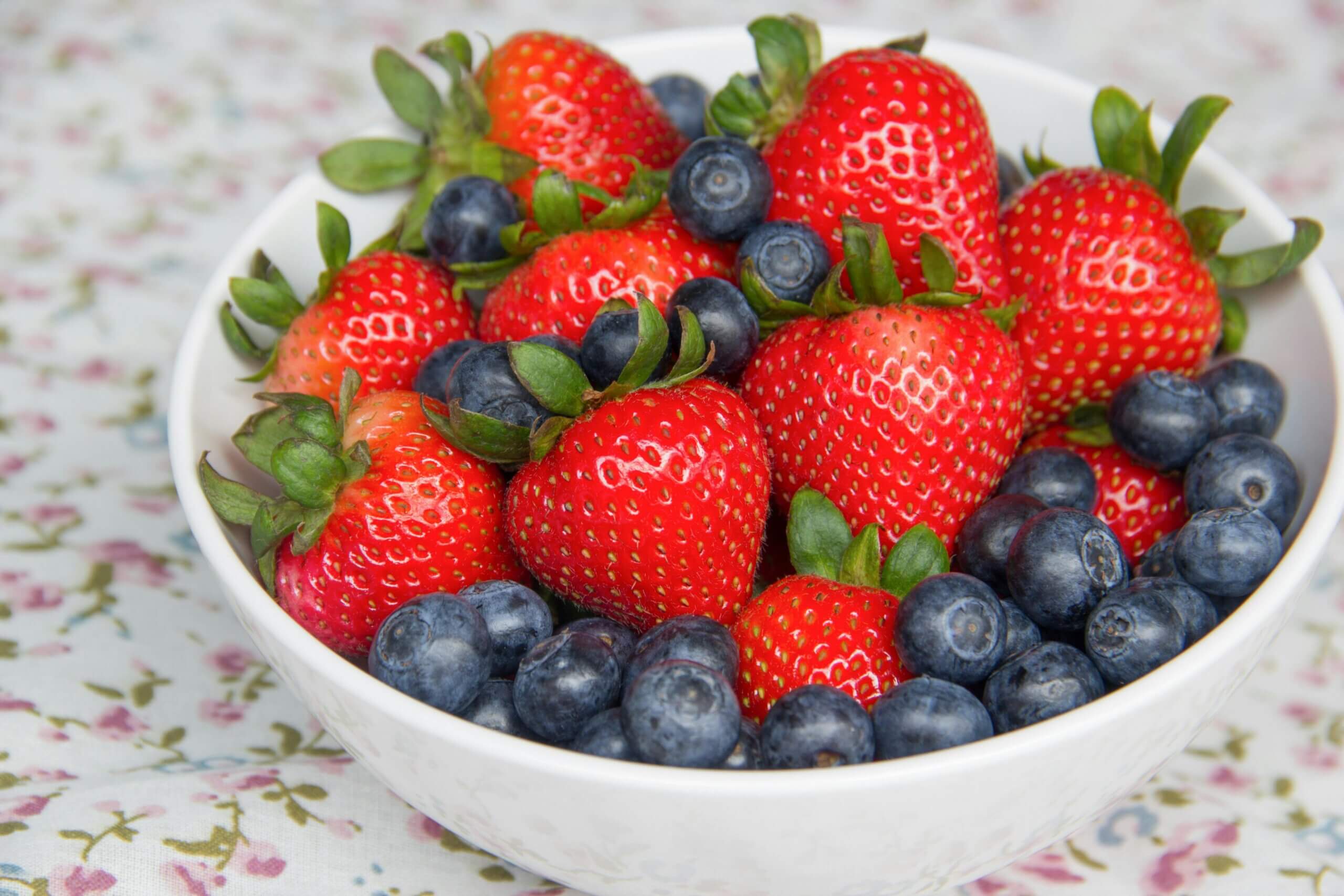 Mangkuk putih berisi stroberi segar dan blueberry di atas taplak meja bermotif bunga