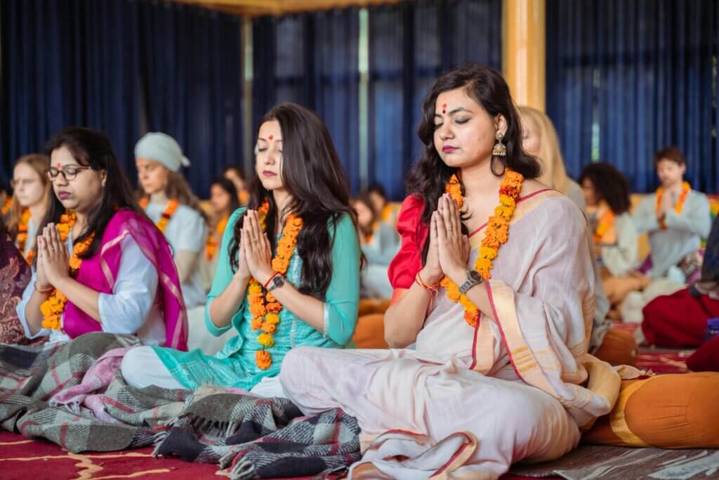 Sekelompok wanita mengenakan karangan bunga marigold bermeditasi dengan tangan dalam posisi berdoa di dalam ruangan
