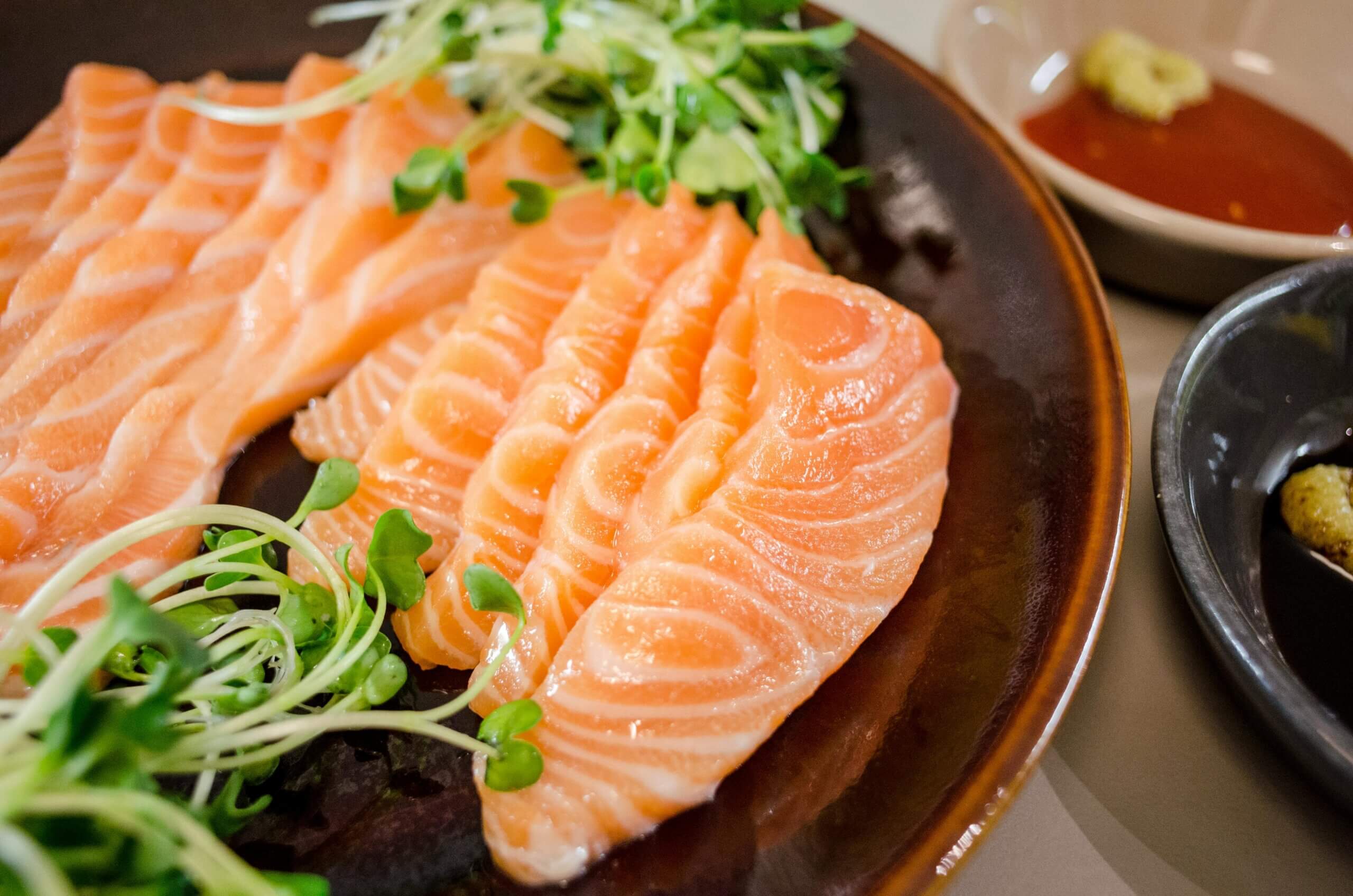 Irisan sashimi salmon segar yang dihias dengan sayuran mikro di piring coklat dengan saus di dekatnya