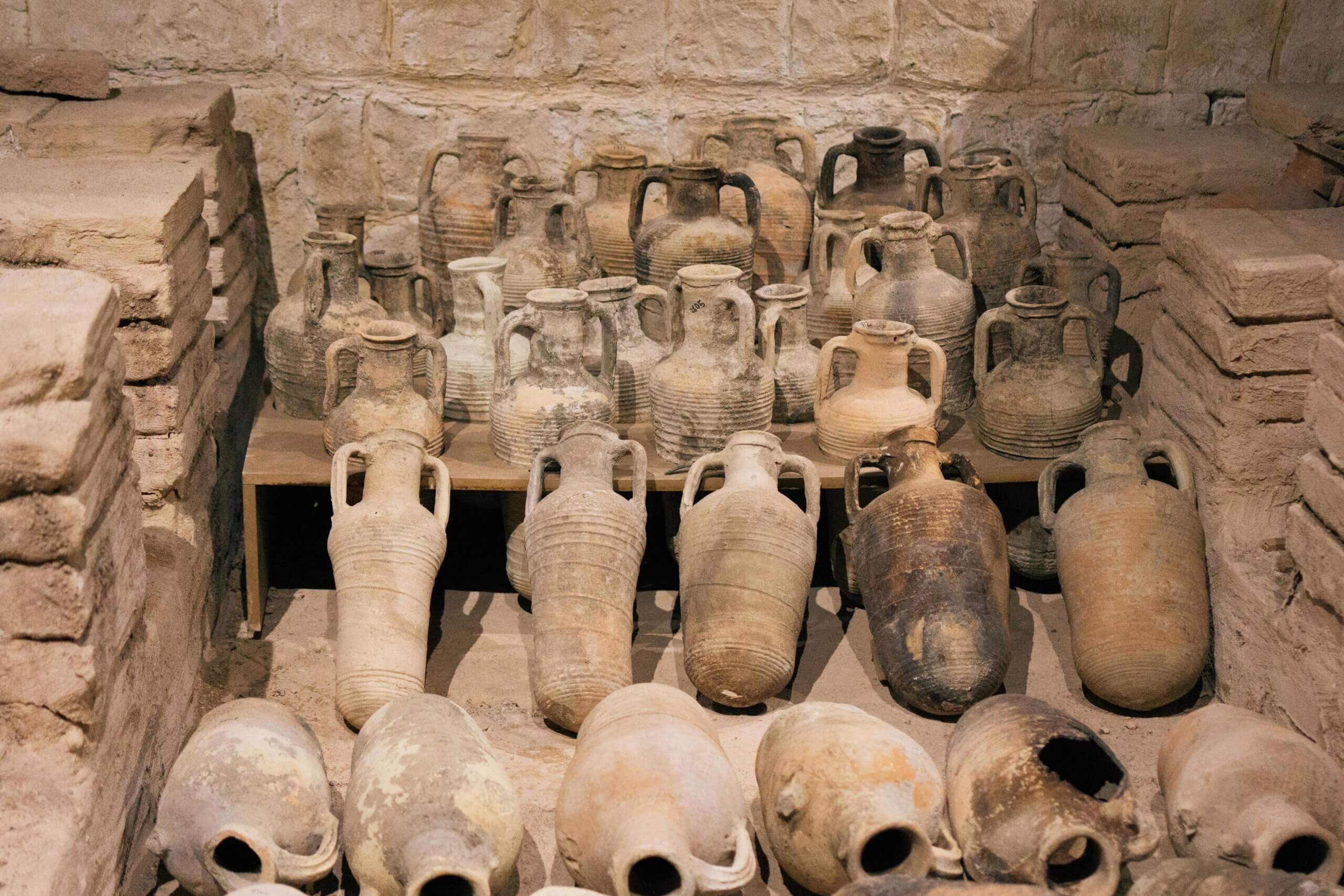 Amphorae tanah liat kuno dan toples tembikar dipajang di situs atau museum arkeologi berdinding batu.