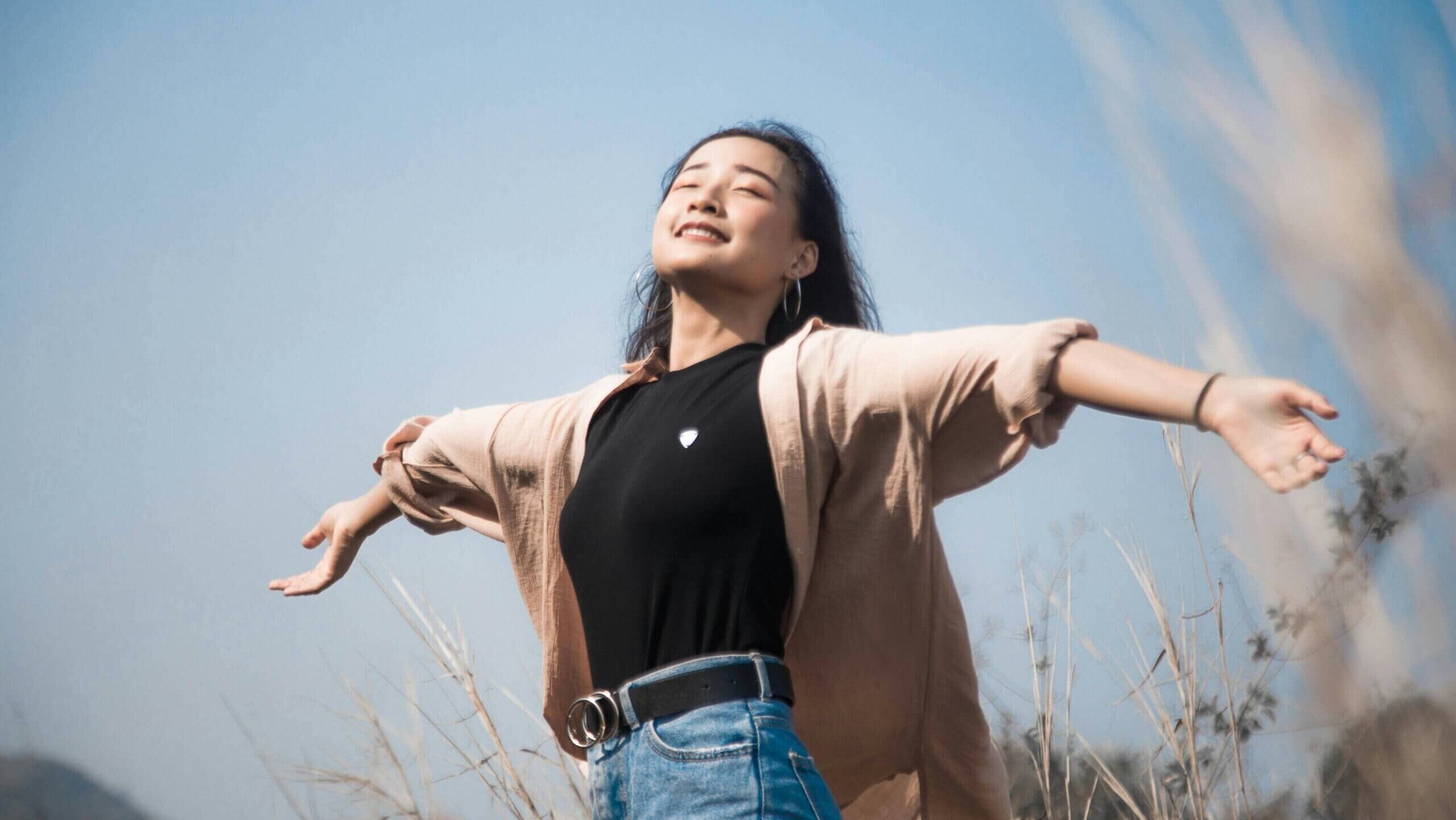 Wanita muda dengan tangan terentang menikmati alam di bawah langit biru cerah di padang rumput tinggi