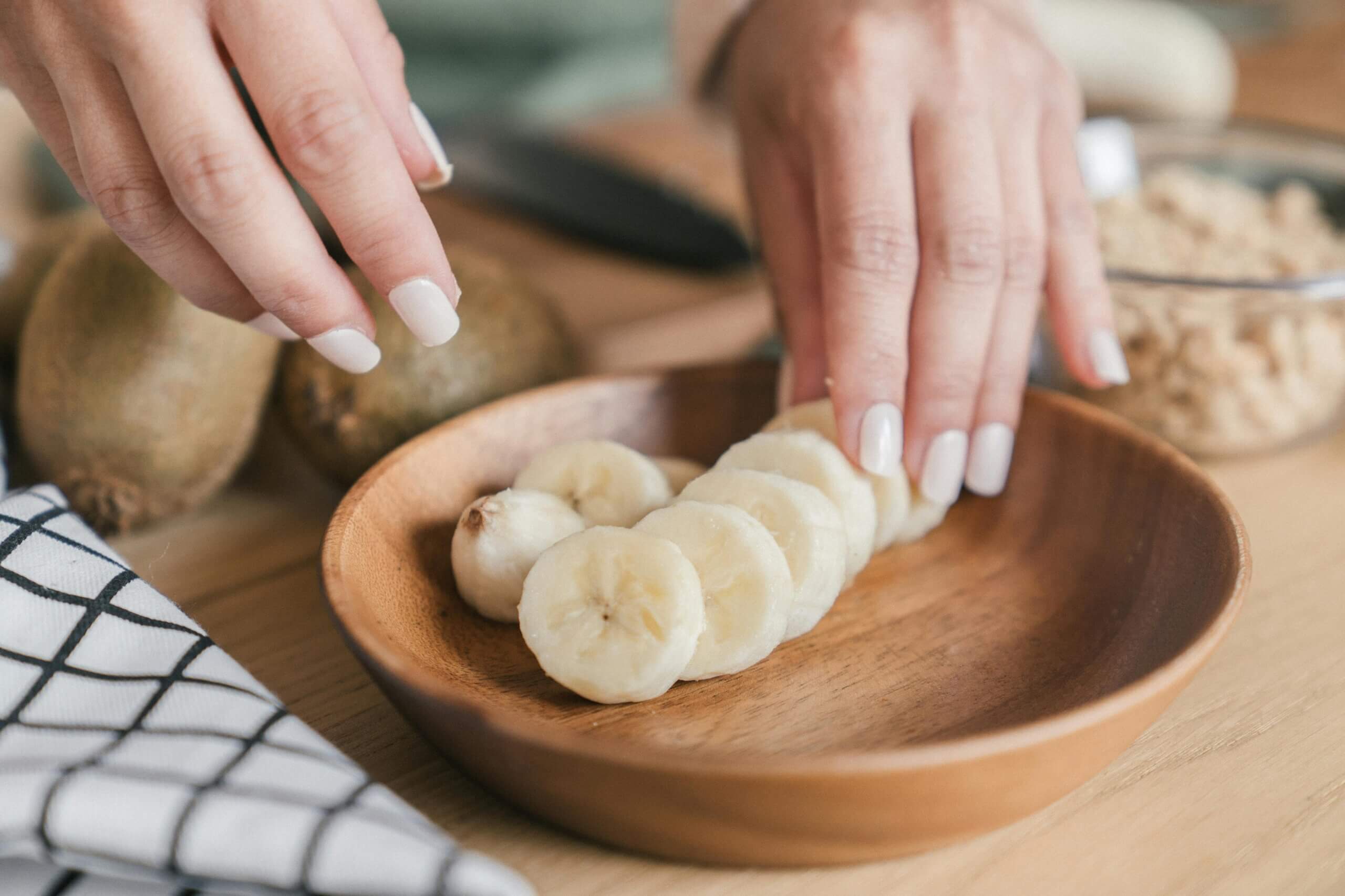 Tangan menata irisan pisang di piring kayu dengan kiwi utuh dan semangkuk sereal di latar belakang