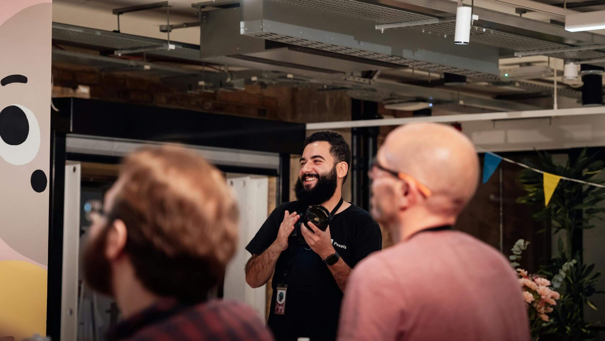 Pria berjanggut memegang kamera dan tersenyum dalam suasana santai di dalam ruangan dengan dua pria kabur di latar depan.