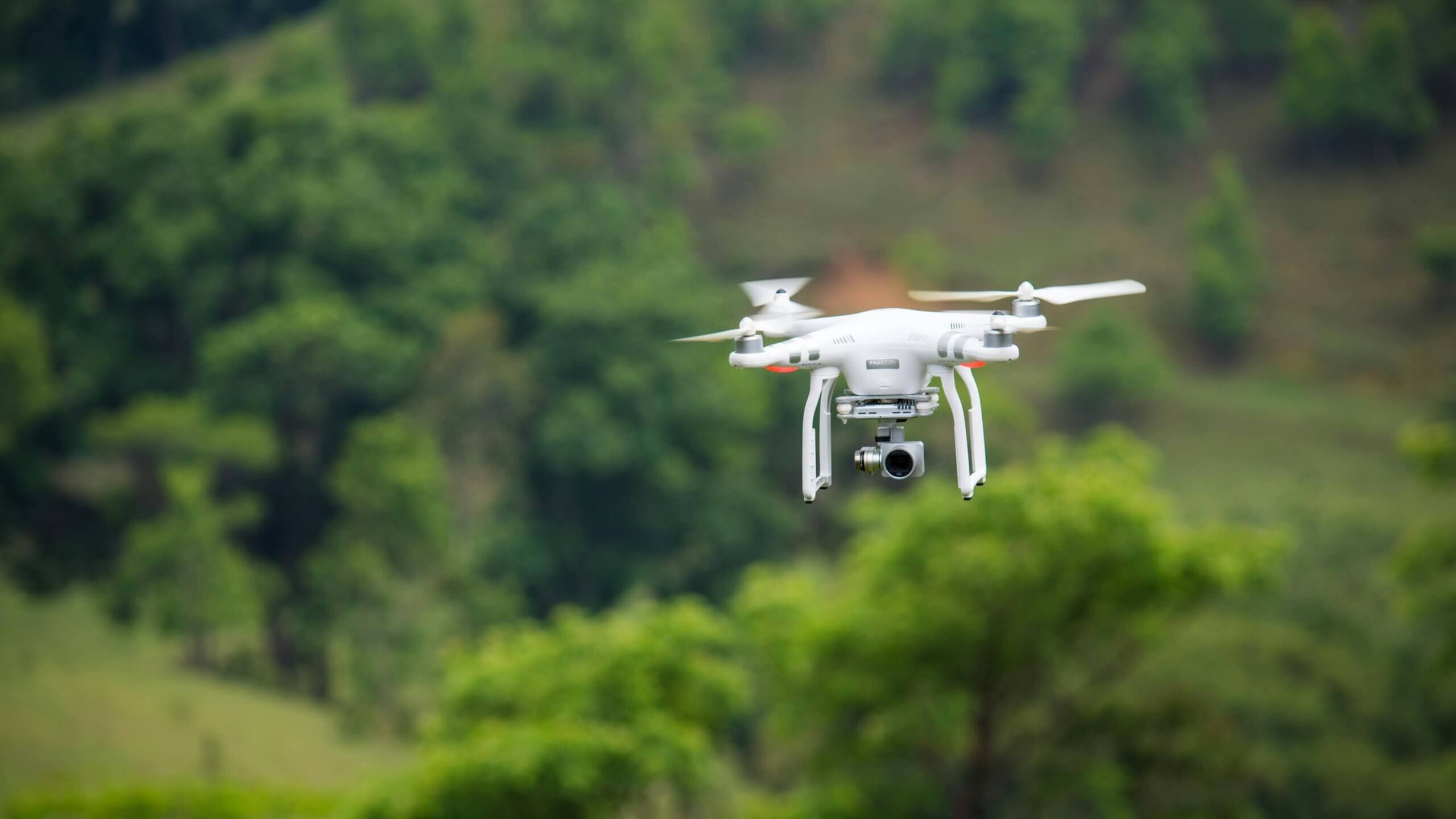 Drone DJI Phantom putih terbang di luar ruangan dengan pepohonan hijau di latar belakang