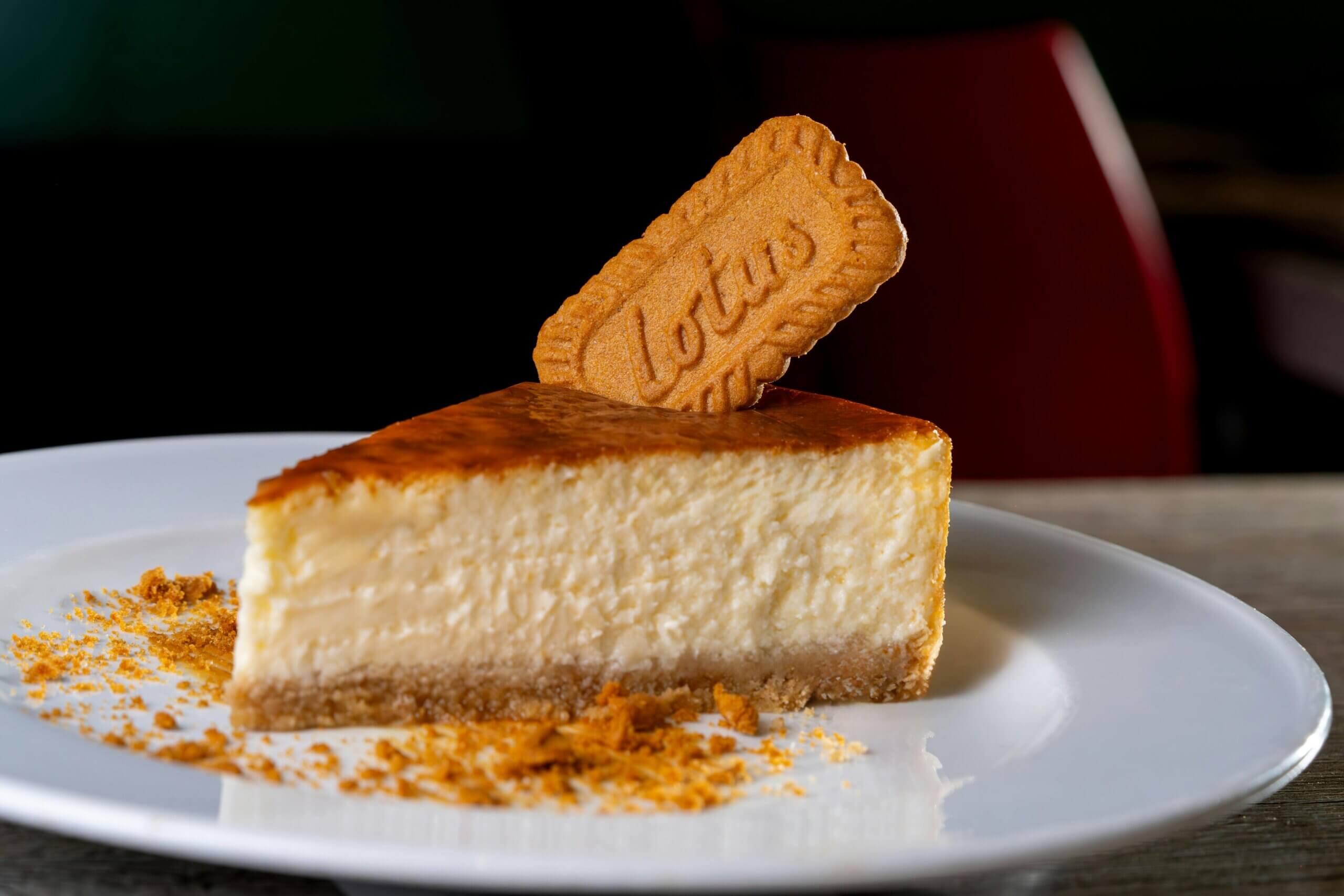 Sepotong kue keju di atasnya diberi kue Lotus Biscoff dan remah-remah di piring putih